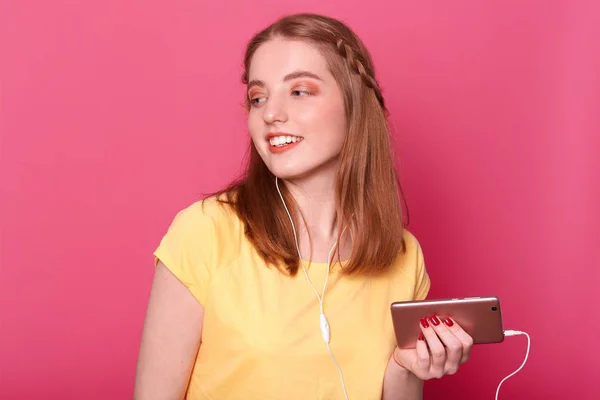 Die fröhliche, unbeschwerte junge Frau hört gerne Musik vom Smartphone, hat Kopfhörer, trägt lässig gelbes T-Shirt, schaut irritiert zur Seite, posiert vor rosa Hintergrund. Unterhaltungskonzept. — Stockfoto