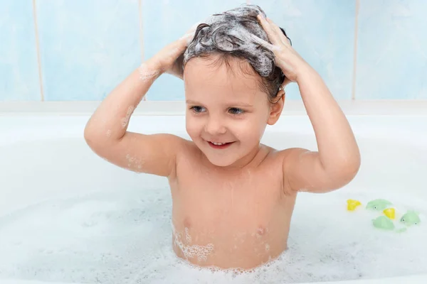 Menina bonito tomando banho, lavar o cabelo com xampu, parece satisfeito beeig na banheira sozinho, brincando com bolhas de espuma, olhando para a câmera, bebê independente gosta de estar em água morna . — Fotografia de Stock