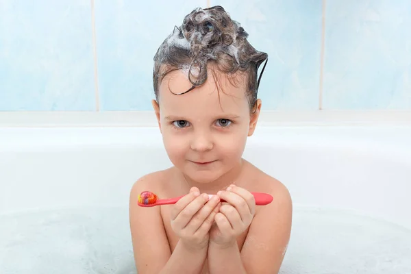 Portret ot aantrekkelijk klein kind kijken camera, houdt rode tandenborstel in handen, klaar voor het tandenpoetsen, geïsoleerd over blauwe muur in badkamer, wordt blij om bad te nemen en gefotografeerd. — Stockfoto