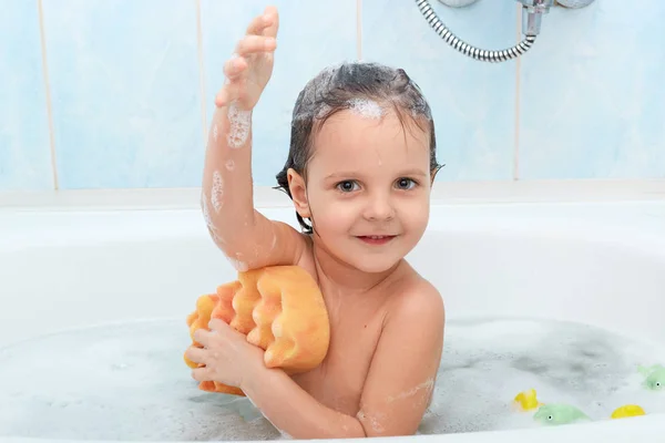 Vrolijke positieve schattig klein kind nemen bad en wassen zichzelf met de gele spons, spreekt aangename emoties, wordt blij om te ontspannen, geïsoleerde op blauwe muur in badkamer. Hygiëne concept. — Stockfoto