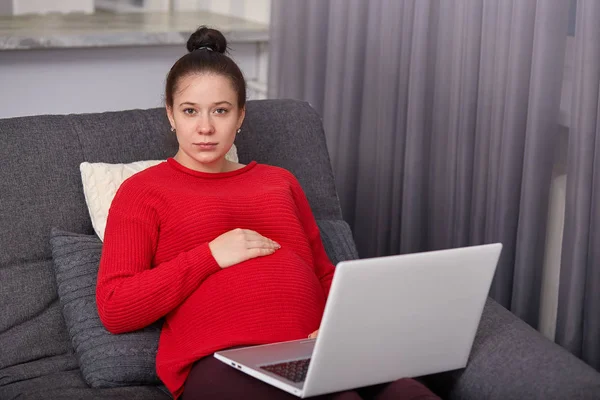 Beautiful pregnant keeps hand on belly, anticipates for baby, looks at camera, sits on comfortable sofa with lap top computer, leans on pillow, dressed in red jumper, has bunch. Pregnancy, technology — Stock Photo, Image