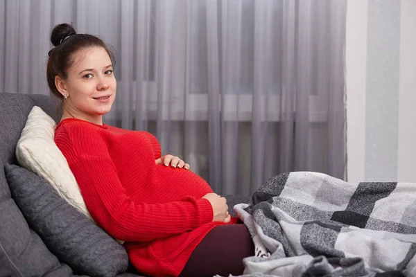 Imagen de la mujer embarazada tranquila lleva suéter rojo, mantiene las manos en el estómago, mira a la cámara, sueños para el bebé, se sienta en el sofá cómodo en la sala de estar, cubre las piernas con. Concepto de embarazo y feminidad . — Foto de Stock