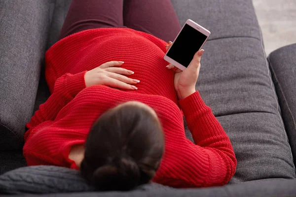 Top view of pregnant female keeps hand on belly, holds mobile phone with blank screen, wears red sweater, with bunch, checking email, lies on comfortable sofa, having rest after going for walk. — Stock Photo, Image