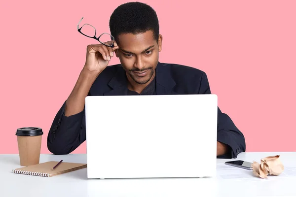 Young black business man with laptop, sits at white table, holds his spectacles in hand, has thoughtful facial expressions, thinks about important contract, poses isolated over rose background.