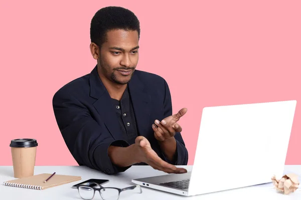 Ratloser junger Geschäftsmann in schwarzer Jacke, zeigt mit beiden Händen auf Laptop-Monitor, sieht Erregung, versteht nicht, was passiert ist, nutzt drahtloses Internet, arbeitet online, verdient Geld im Büro. — Stockfoto