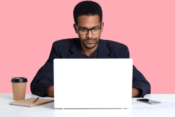 Close up retrato de homem de pele escura veste terno preto, trabalha on-line com lap top, freelancer afro-americano senta-se à mesa branca na frente do computador portátil aberto, isolado sobre fundo rosa . — Fotografia de Stock