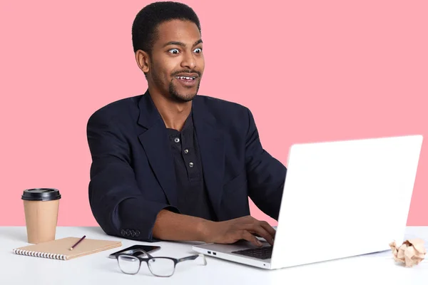 Retrato interno de jovem macho preto surpreso sentado à mesa, trabalha com regaço, olha com espanto no monitor. Bonito tipo a escrever no teclado, expressa choque com a cara. Conceito de trabalho online — Fotografia de Stock