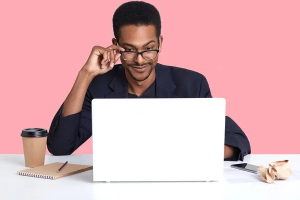 Retrato de homem de pele escura trabalha com laptop. Bonito macho preto se senta na mesa usa terno, toca seus óculos com a mão, olha concentrado no monitor. Conceito de trabalho e tecnologia online — Fotografia de Stock