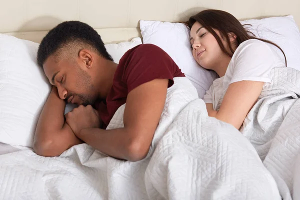 Closeup of couple sleeping in bed, having rest after hard day, dressed t shirts, lie with closed eyes, try to fall asleep, husband offended by his wife and turned his back. Relation and people concept — Stock Photo, Image