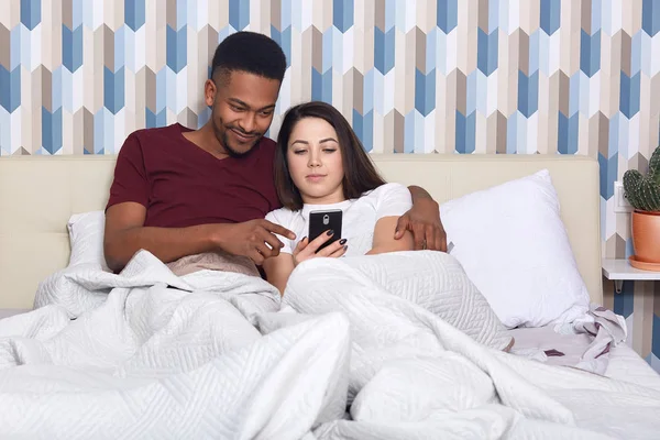 Young couple lying in bed, female iholding mobile phone, husband hugs his wife, watching film online, having rest together, isolated on colourful wall. People, relation, resting, comunication concept. — Stock Photo, Image