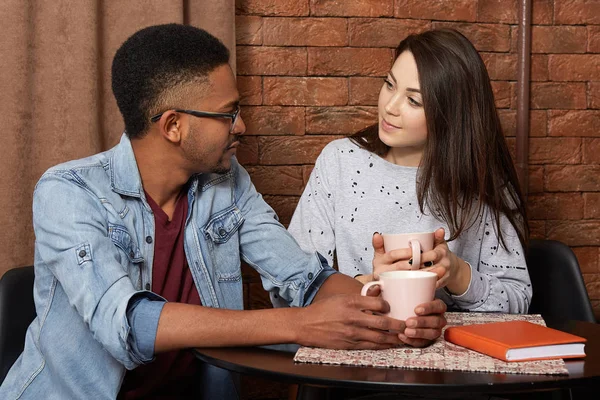 Romantic relaxed couple sit at table in cafe, look at each other, hold cups of coffee or tea in hands, being of different race, enjoy togetherness, spend weekend together. People, relationship concept