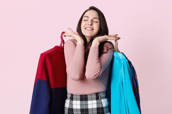 Aantrekkelijke brunette vrouw draagt roze shirt en rok gecontroleerd heeft verheugd expressie, staat met gesloten ogen, ziet er zeer gelukkig, houdt hangers met kleren. Mensen, winkelen en geluk concept. — Stockfoto