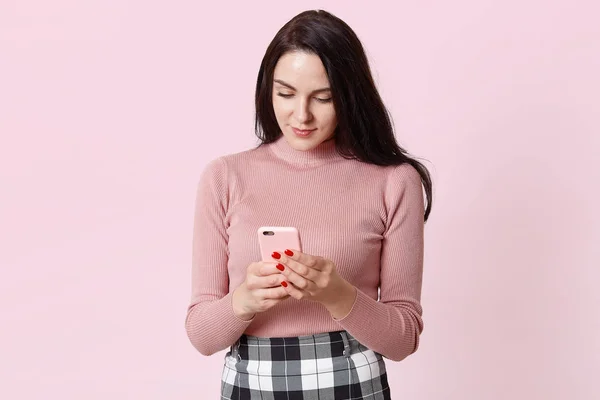 Внутренний снимок концентрированной нежной молодой женщины держит в руке мобильный телефон, читает сообщение от бойфренда, проверяет женщину, позирует в фотостудии, изолирован на розовой стене, копирует пространство для рекламы . — стоковое фото
