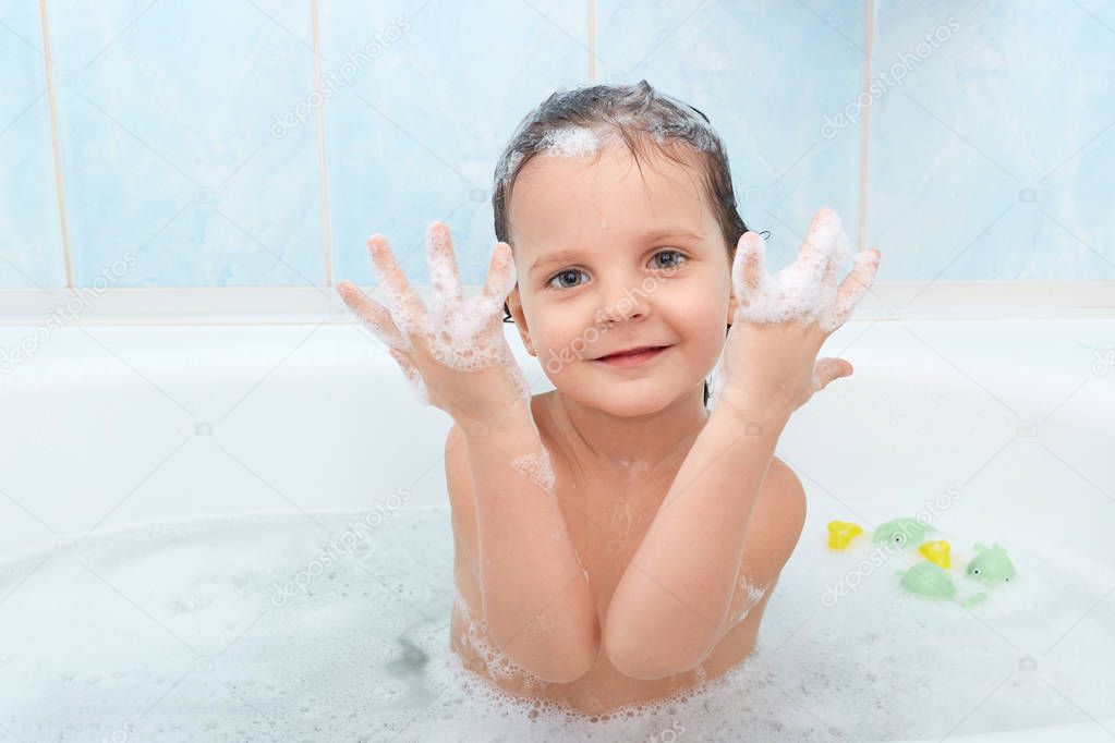 Small adorable child has funny expression, taking bath with plesure, plaing in hot water and shows hands with foam, poses in bathroom against blue wall. Playful little kid glad to be photographed.