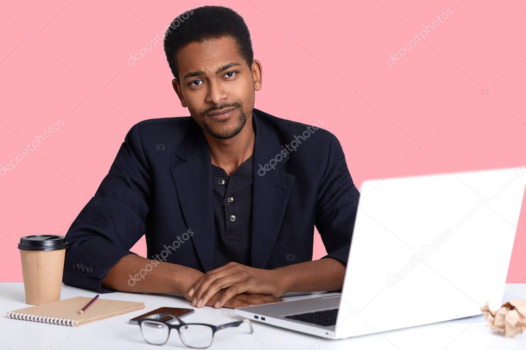 Photo of handsome african american male taking brake from working with laptop computer, looks axuasted at camera, has upset facial expressions isolated over pink background. People concept.