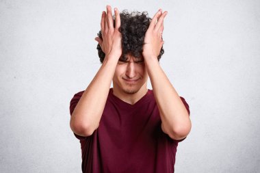 Kıvırcık saçlı çekici erkek casual bordo t gömlek giyer, mutsuz expresions vardır, beyaz arka plan üzerinde izole eliyle başını dokunur. Kopya alanı reklam veya promosyon metni için.