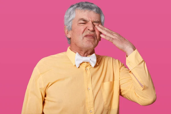 Close up ritratto di bel grigio capelli uomo maturo tiene le dita sugli occhi e strofinando, indossa camicia gialla in un tono e papillon bianco, vuole addormentarsi, isolato su sfondo studio . — Foto Stock