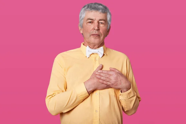 Handsome middle age hoary senior man wearing yellow shirt and white bow tie isolated over pink background smiling , stands with hands on chest, looks at camera and shows grateful gesture on face. — Stock Photo, Image