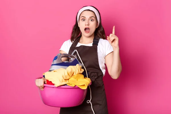 Housewife holds basin of fresh clothes, stand with open mouth, has shocked facial expressions, resdy to iron, remember some important chores, isolated over pink background. Household, domestic concept — Stock Photo, Image