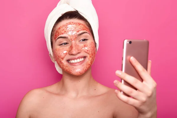 Blij blij meisje met zachte huid maakt selfie tijdens het deinende spa procedure, draagt witte handdoek, heeft blij look poses glimlachend geïsoleerd op roze achtergrond. Mensen, de schoonheid en de huid zorg concept — Stockfoto