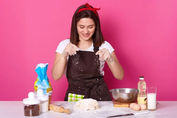 Young woman baker in kitchen, sprinkling white flour on dough, baking delicious coockies, likes homemade pastry, posing isolated over pink background. Copy space for your advertismant or promotion.