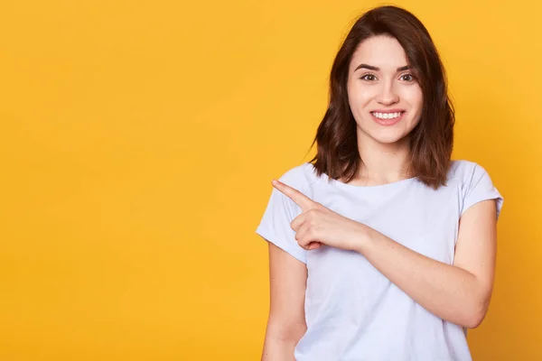 Retrato de bonito encantador menina alegre apontando para o lado com o dedo indicador, veste t-shirt casual branco, tem cabelo escuro, isolado sobre fundo amarelo. Copiar espaço para o seu texto de promoção ou publicidade — Fotografia de Stock