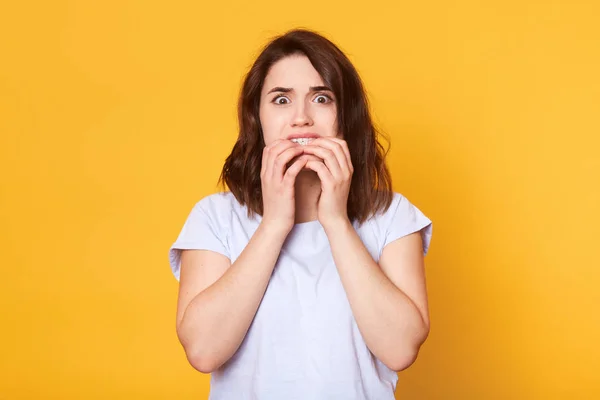 Retrato de chica morena asustada muerde las uñas de los dedos, tiene espantosa necesidad de expresión facial, temores de algo, lleva puesta una camiseta blanca casual, modelos sobre fondo amarillo. Concepto de sentimientos negativos — Foto de Stock