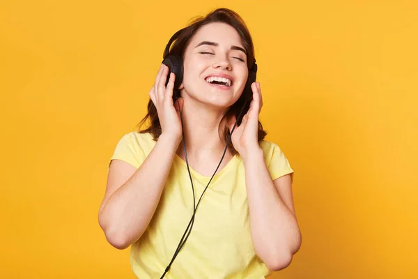 Hermosa mujer escuchando música sobre fondo amarillo. Encantadora dama posando con los ojos cerrados, disfruta de escuchar música favorita, mantiene las manos en los auriculares, canta y se relaja. Concepto de estilo de vida . — Foto de Stock