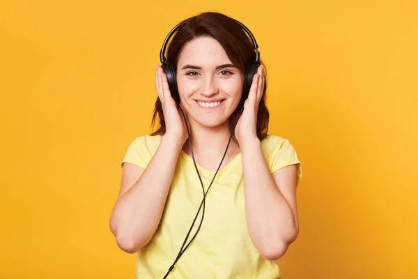 Image of young happy woman with headphones listening to music isolated over yellow background wall, being in good mood, stands smiling and looks directly at camera. Copy space for advertisment. — Stock Photo, Image