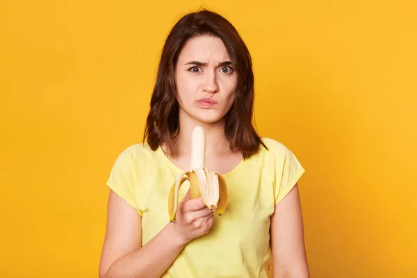 Foto de mujer de aspecto agradable en ropa casual, lista para comer plátano fresco, no le gusta esta fruta, tiene una expresión facial desagradable, posa sobre un fondo amarillo. Nutrición saludable — Foto de Stock