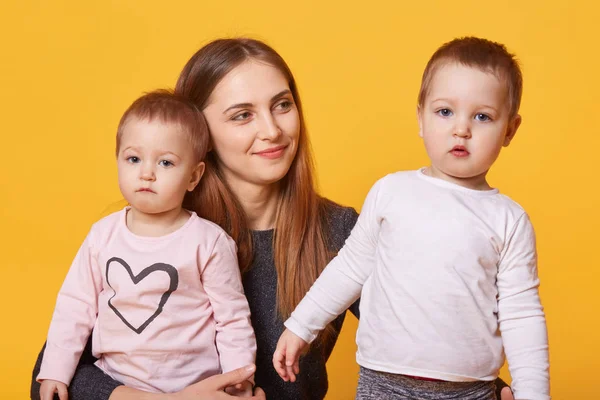 Mujer feliz, madre y sus hijos gemelos posa en un estudio fotográfico aislado sobre un fondo amarillo. Mami mira a sus bebés con gran amor, abraza a sus doncellas. Copia el espacio para tu anuncio . — Foto de Stock