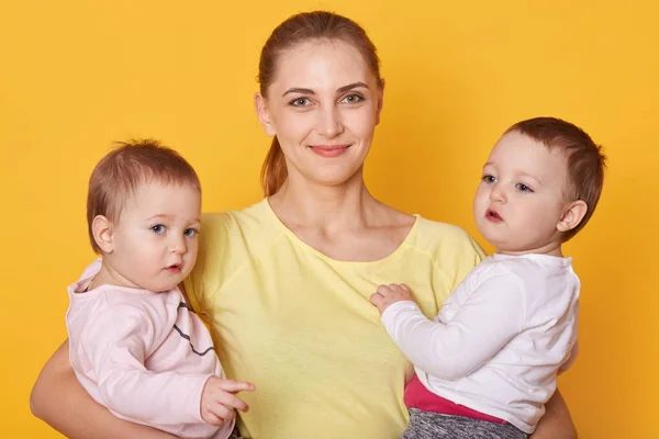 Image de mère avec des enfants, deux filles en vêtements décontractés, belle jeune femme avec des petits jumeaux debout dans un studio photo isolé sur fond jaune. Les filles étant intéressées à poser avec maman . — Photo