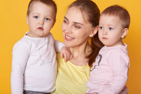 Portrait of young mother holding her little twins, cute active infants being interested to be photografed, look asides, mother looks with love on her adorable daughters. happy family concept. — Stock Photo, Image
