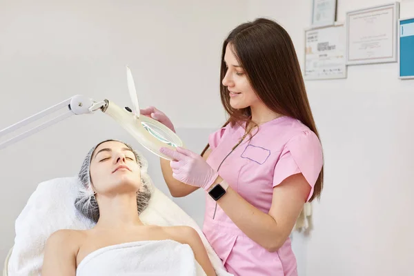 Młoda kobieta chce oczyszczanie twarzy, badane przez specjalistę beutician pod lampą, klient siedzi na couh podczas leczenia twarzy klinika urody. Pielęgnacja skóry, kosmetyki, koncepcja medycyna aethetic. — Zdjęcie stockowe