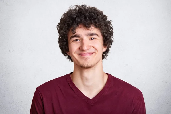 Headshot of pleased hipster guy dressed in maroon t shirt, enjoys spare time, has dark curly hair, models over white background. People, youth and lifestyle concept. Copyspace for advertisement. — Stock Photo, Image