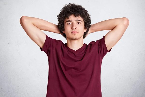 Afbeelding van man met krullend haar heeft zijn handen achter zijn hoofd, ziet er serieus direct bij camera, draagt casual kastanjebruine t shirt geïsoleerd op witte achtergrond. Ruimte voor uw advertentie kopiëren. — Stockfoto