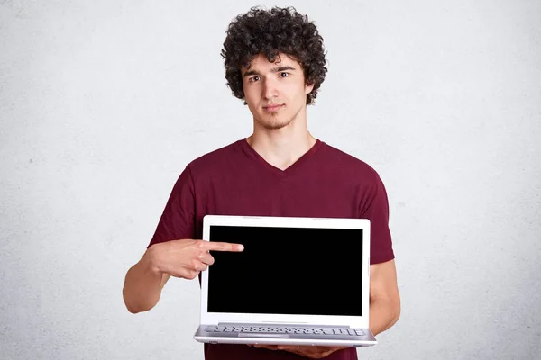 カジュアルなあずき色の t シャツ、ラップトップ コンピューターが付いている立場で若いハンサムな男の画像は、白い壁、広告コンテンツやプロモーションの空き領域に分離された空の画面で前面の指. — ストック写真