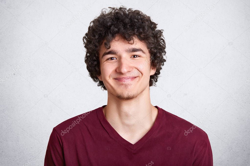 Headshot of pleased hipster guy dressed in maroon t shirt, enjoys spare time, has dark curly hair, models over white background. People, youth and lifestyle concept. Copyspace for advertisement.