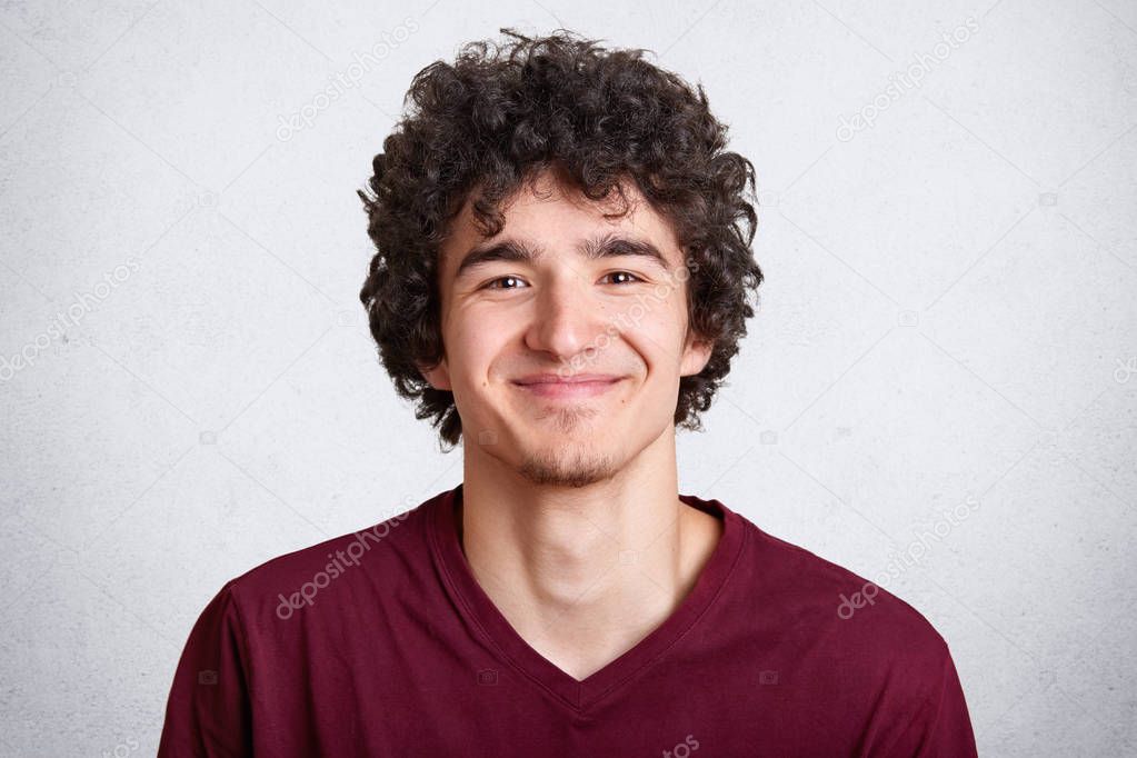 Photo of handsome unshaven guy looks with pleasent expression directly at camera, wears fashionable maroon t shirt, stands against white studio wall. People, lifestyle and fashion concept. Copy space.