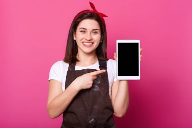 Happy young woman showing tablet computer screen, copy space for your advertisment or promotion text, isolated over pink background, looks at camera, wears white t shirt, brown apron and red hair band