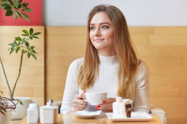Portrait of good-looking young female sitting in cafe during lunch. Adorable blond haired lady holds cup of hot drink in her hands, wears white dress, looks out of window dreamy, enjoys her rest.