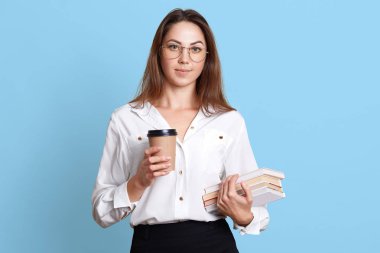 Mavi arka planda izole, trendy gözlük sert çalışan ciddi genç bayan portresi. Avrupa esmer model kitap demet ve kahve bir fincan kağıt tutar. Reklam için alanı Kopyala.