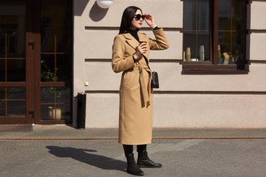 Genç hoş Blackhaired kadın mesafe görünüyor, uzun yürüyüş için daha iyi yol seçmek için çalışır. Moda uzun boylu Bayan deri çanta ve siyah güneş gözlüğü ile uzun bej şık ceket giyer.