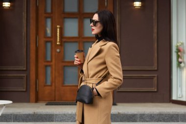 Charming gizemli kadın dışarıda duruyor, bej ceket giyer, deri siyah çanta, şık güneş gözlüğü, cebinde elini tutar, diğer taraftan kahve bir kağıt fincan var, başka bir şekilde görünüyor.