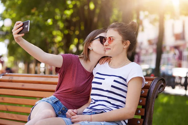 Mutlu samimi iki kız parkta oturan ahşap tezgah üzerinde selfie yapmak. Fotoğraf çekerken neşeli genç kız sağ yanak onu en iyi arkadaşı öpüyor. Arkadaşlar fotoğrafları görmek için sabırsızlanıyoruz. — Stok fotoğraf