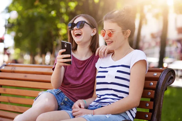 Leales amigos felices se sienten bien cerca el uno del otro en el parque. Hermosas chicas alegres leen divertida historia impredecible en el banco estalló riendo en voz alta y descansar juntos en el fin de semana de verano . — Foto de Stock