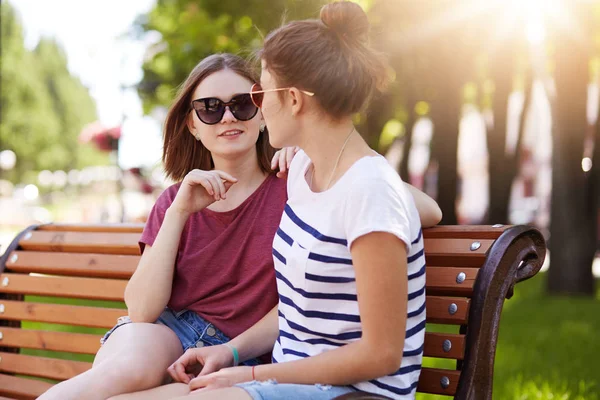 Agradable chicas habladoras tienen buena conversación sobre los últimos acontecimientos en sus vidas. Adorables amigos se sientan en un banco de madera en el parque local y disfrutan de un clima soleado, cálido y de verano. Concepto de amistad . — Foto de Stock