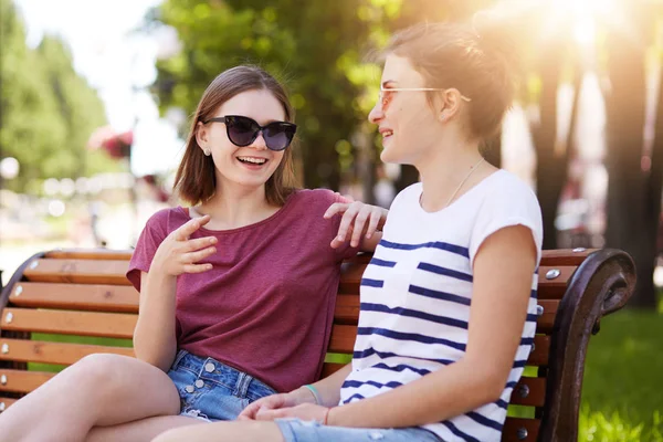 Dos niñas alegres relajarse juntos en el parque local sentado en un nuevo banco de madera, hacer bromas, chismes, recordar viejos momentos divertidos entre sí. Chicas lindas atractivas no pueden dejar de reír. Concepto de amistad . — Foto de Stock