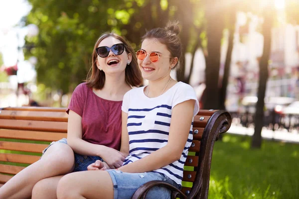 Un retrato de dos jóvenes amigos atractivos que descansan en un banco de madera en el parque local después de un largo paseo. Las chicas que ríen tienen mucha energía para continuar su diversión. Concepto de relaciones de amistad . — Foto de Stock