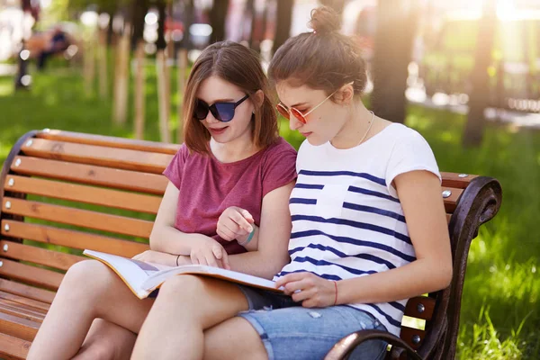 Promyšlené dvě dívky nosí neformální oblečení, sedí na dřevěné lavičce v parku a hovoří o novinkách popsaných v nejnovějším časopise, čtou legrační věci. Přátelě přátelé čtou a mluví spolu. — Stock fotografie
